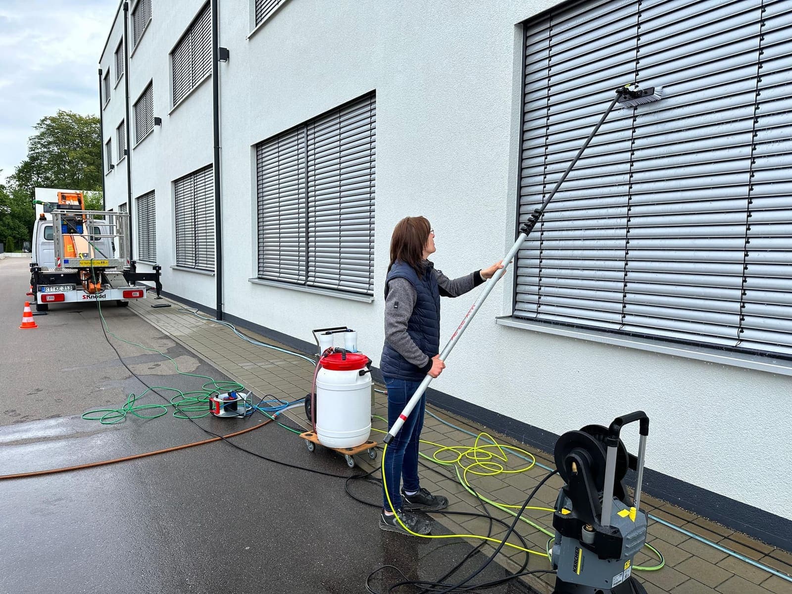 Mitarbeiterin reinigt Fensterjalousien am Gewerbegebäude mit Teleskopstange, Heißwasser- und Osmose-Gerät im Vordergrund