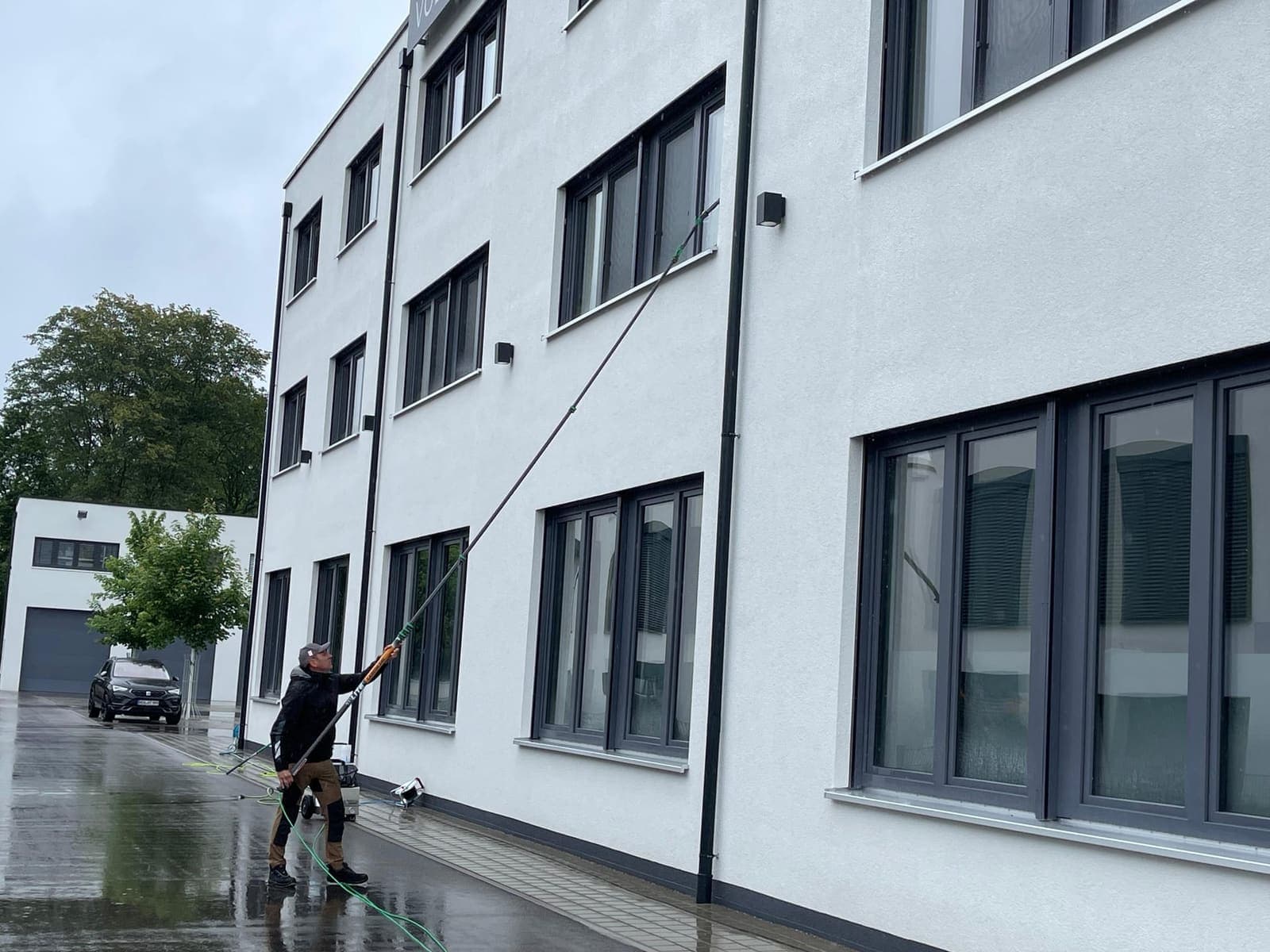Mitarbeiter reinigt Fenster eines dreistöckigen Gewerbegebäudes mit Teleskopstange bei Regen